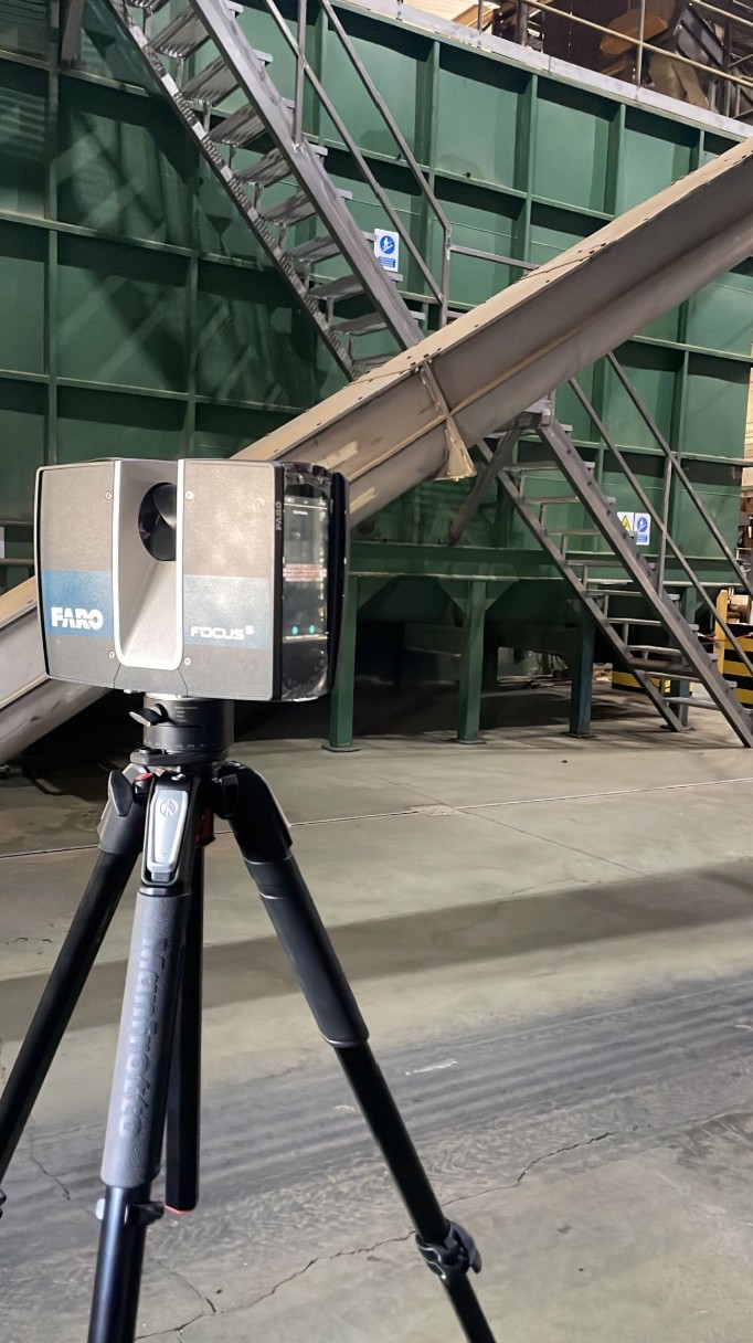 Escáner láser 3D sobre trípode en una planta de tratamiento de residuos, con una cinta transportadora, una estructura metálica y escaleras al fondo
